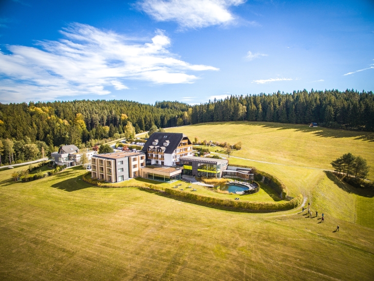 Luftaufnahme eines modernen Hotels in einer ländlichen Umgebung, umgeben von Wäldern und Wiesen.