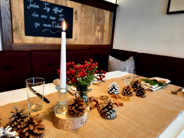 Gedeckter Ecktisch mit rustikalem Tischläufer, einer Kerze und Dekoelementen aus Zapfen und getrockneten Früchten, an der Wand dahinter hängt eine Tafel mit einem handgeschriebenen Spruch