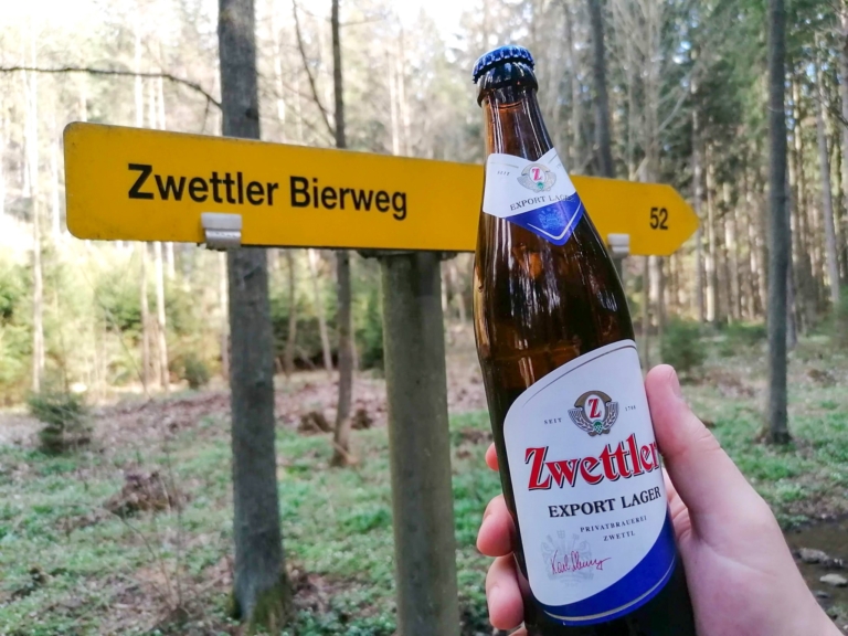 Eine Hand hält eine Flasche Zwettler Bier vor dem Wanderwegweiser zum Zwettler Bierweg in einem Wald